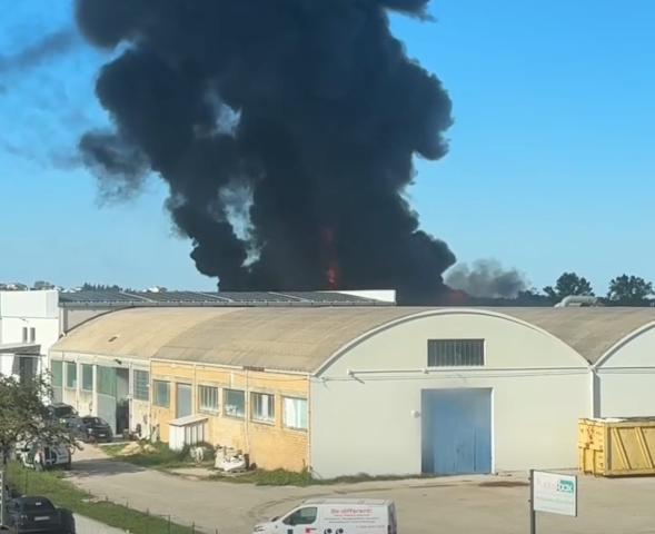 Grosso incendio in un'area industriale, con fumo nero denso che si alza da un edificio. Si vedono capannoni industriali in primo piano, mentre le fiamme sono visibili tra il fumo sullo sfondo.