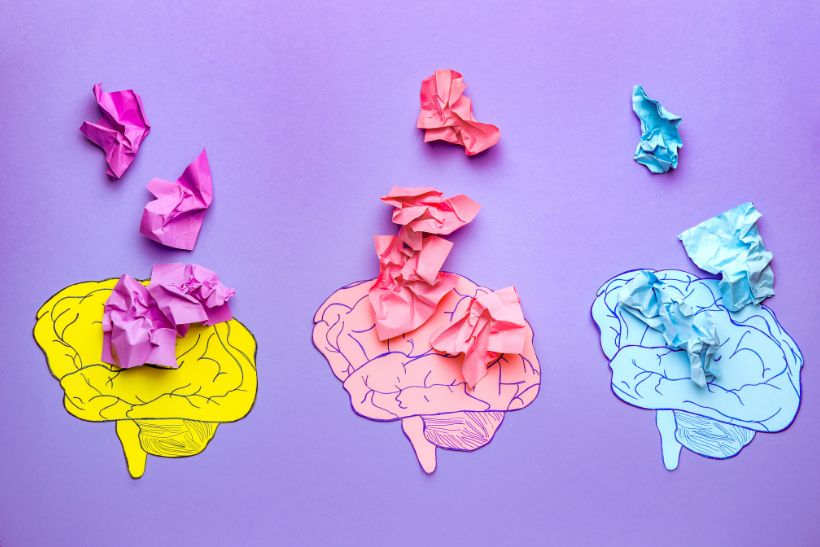 L'immagine raffigura tre cervelli stilizzati su sfondo viola, colorati in giallo, rosa e blu. Su ciascuno ci sono pezzi di carta accartocciati che corrispondono al colore del cervello, simboleggiando probabilmente concetti legati alla creatività - idee