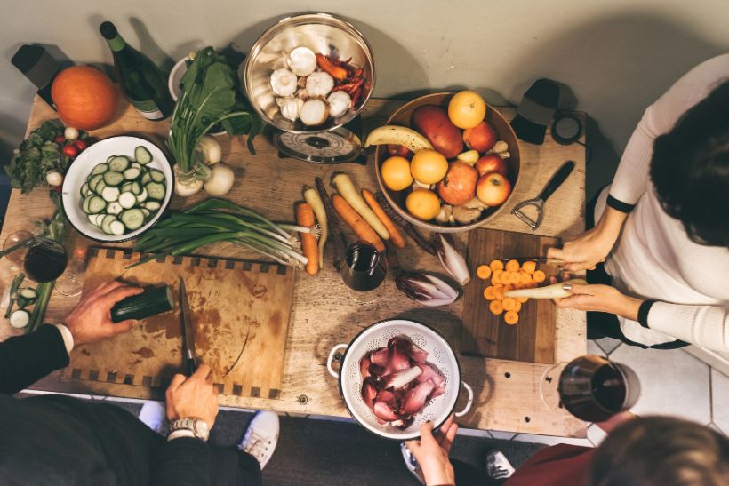 L'immagine mostra tre persone che preparano cibo su un tavolo di legno, tagliando verdure come cetrioli e carote. Il tavolo è ricco di ingredienti freschi, tra cui frutta, aglio e verdure a foglia verde, con bicchieri di vino a fianco.