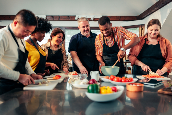 L'immagine mostra un gruppo di persone di diverse origini che partecipano a una lezione di cucina. Tutti indossano grembiuli e sono impegnati a tagliare verdure o mescolare ingredienti. L'atmosfera è conviviale e rilassata.