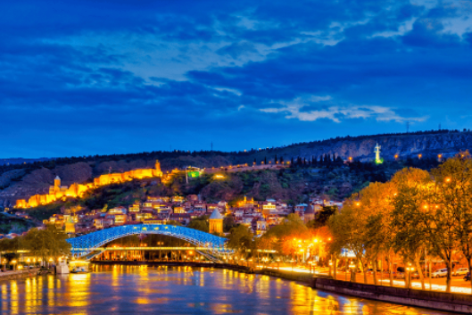 Panorama serale di una città illuminata. In primo piano un fiume con riflessi delle luci circostanti e un ponte moderno illuminato di blu. Sullo sfondo, una collina con edifici e mura storiche illuminate di arancione e una statua ben visibile in cima