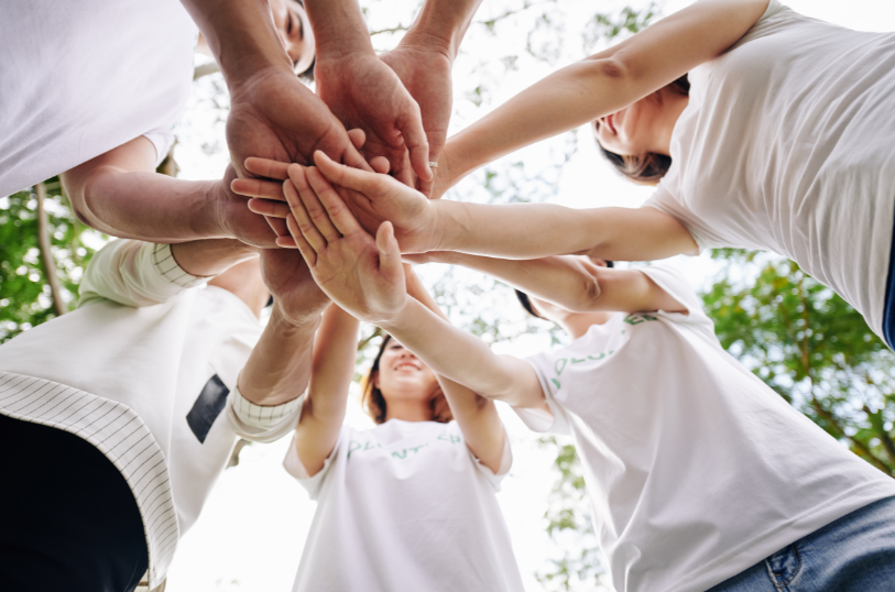 L'immagine mostra un gruppo di persone che uniscono le mani al centro, simbolo di collaborazione e lavoro di squadra. Gli individui indossano magliette bianche, e la scena è all'aperto, suggerendo un contesto naturale o di volontariato.