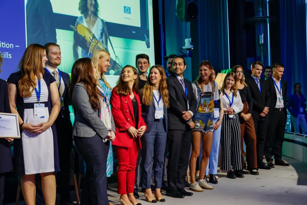 L'immagine mostra un gruppo di giovani in abbigliamento formale su un palco durante un evento ufficiale dell'UE, sorridenti e con badge identificativi. Sullo sfondo, un grande schermo proietta un'immagine con il logo dell'Unione Europea